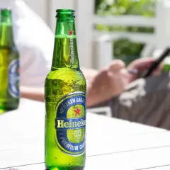 Orzeźwiająca butelka Heineken 0.0 procent, idealna na letni relaks. W tle osoba korzystająca z telefonu.
