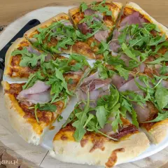 Pyszne pomysły na pizzę: cienkie ciasto, sos pomidorowy, ser, szynka i rukola. Obok leży nóż do pizzy.