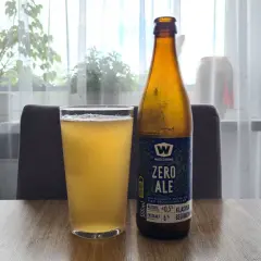 Szklanka i butelka piwa zero na drewnianym stole. Za nimi okno z firanką i roślinami.