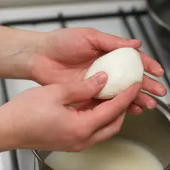 Dłonie trzymają kulkę mozzarelli nad garnkiem z mlekiem, sprawdzając, czy mozzarella w kulce się rozpuszcza.