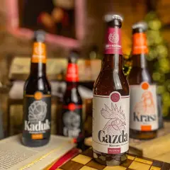 Piwo z Pomorza: butelki "Kaduk", "Gazda" i "Krasa" stoją na stole obok otwartej książki i szachownicy.
