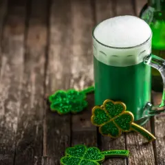 Zielone piwo irlandzkie z pianką, butelka i błyszczące koniczynki na drewnianym stole.