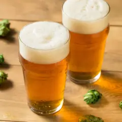 Dwa szklanki orzeźwiającego hazy IPA na drewnianym blacie, otoczone świeżymi szyszkami chmielu.