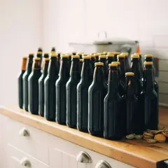 Rząd ciemnych butelek z piwem jęczmiennym na drewnianym blacie. Kilka kapsli leży obok.