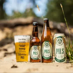 Szklanka, butelka i puszka piwa lager "Pils" od Trzech Kumpli na piasku, wśród traw.