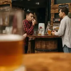 Dwóch mężczyzn rozmawia przy barze, zastanawiając się, ile węglowodanów ma piwo. Na stole stoją kufle i beczka.
