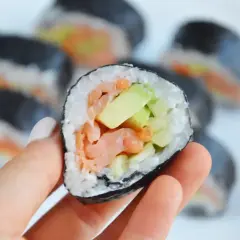Ręka trzyma kawałek sushi z łososiem i awokado w środku. Na talerzu widać więcej takich roladek.