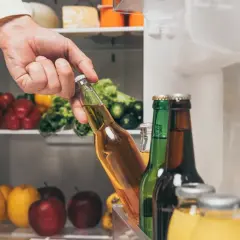 Ręka wyjmuje butelkę piwa z lodówki, gdzie obok stoją inne butelki i owoce. Czy piwo się psuje? Nie, jeśli jest schłodzone!