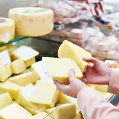 Ręce trzymają kawałki żółtego sera, zastanawiając się, jaki ser ma najmniej kalorii. W tle widać półki pełne różnych serów.