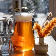 Dzbanek orzeźwiającego helles bier z pianką obok szklanki i ozdobnej trawy.