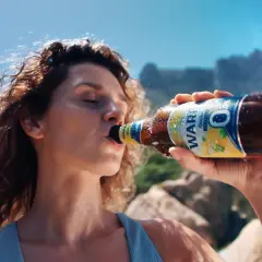 Kobieta pije piwo Żywiec 0%. Na plaży, w tle góry i palmy.