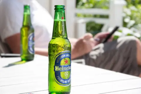 Orzeźwiająca butelka Heineken 0.0 procent, idealna na letni relaks. W tle osoba korzystająca z telefonu.