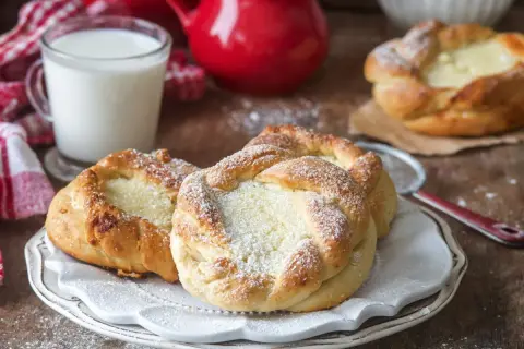 Puszyste drożdżówki z serem, posypane cukrem pudrem, obok szklanka mleka i czerwone dzbanki. Idealne na śniadanie.