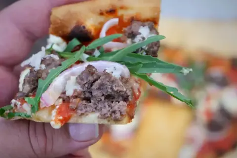 Kawałek pysznej pizzy 30 cm z mielonym mięsem, rukolą i czerwoną cebulą, trzymany w dłoni.