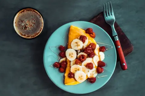 Słodki omlet z bananami i malinami, polany syropem, obok filiżanka kawy. Idealny na słodki początek dnia.