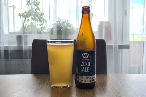 Szklanka i butelka piwa zero na drewnianym stole. Za nimi okno z firanką i roślinami.