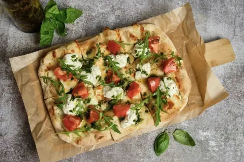 Pyszna pizza wege z pomidorami, mozzarellą i rukolą, polana zielonym pesto. Idealna na lekki posiłek.