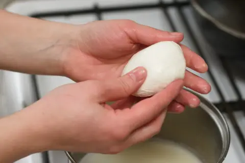 Dłonie trzymają kulkę mozzarelli nad garnkiem z mlekiem, sprawdzając, czy mozzarella w kulce się rozpuszcza.