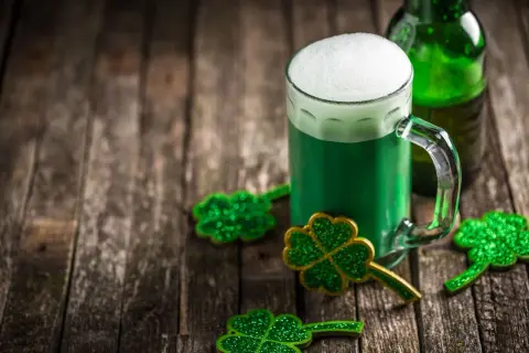 Zielone piwo irlandzkie z pianką, butelka i błyszczące koniczynki na drewnianym stole.