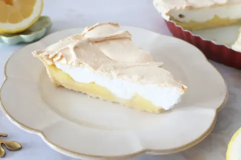 Pyszna tarta cytrynowa z puszystą bezą na wierzchu, podana na białym talerzu. Obok leży połówka cytryny.