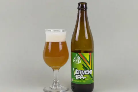 Szklanka z bursztynowym piwem Vermont IPA i butelka tego piwa. Idealne neipa piwo na wieczór.