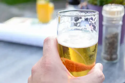 Dłoń trzymająca szklankę orzeźwiającego piwa dietetycznego. W tle widać pomarańczowy napój i młynki do przypraw.