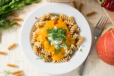 Makaron fusilli z kremowym sosem z **hokkaido dyni**, udekorowany koperkiem i chili. Obok talerza leży cała dynia.