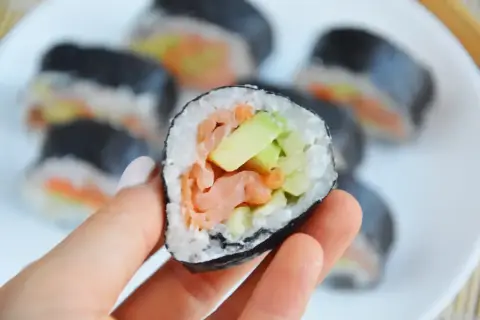 Ręka trzyma kawałek sushi z łososiem i awokado w środku. Na talerzu widać więcej takich roladek.