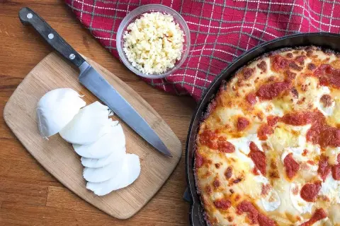 Idealny jaki ser do pizzy? Na desce świeża mozzarella, obok nóż i miseczka z tartym serem. Obok pyszna, gorąca pizza.