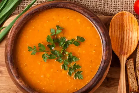 Aksamitna zupa pomidorowa w drewnianej misce, udekorowana natką pietruszki. Obok łyżka i kromki chleba.