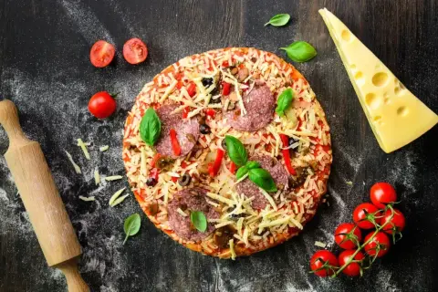 Gotowa pizza z salami i serem, obok wałek, pomidorki i ser. Idealna do domowego wypieku, zastanawiasz się, jaka mąka do pizzy domowej będzie najlepsza?