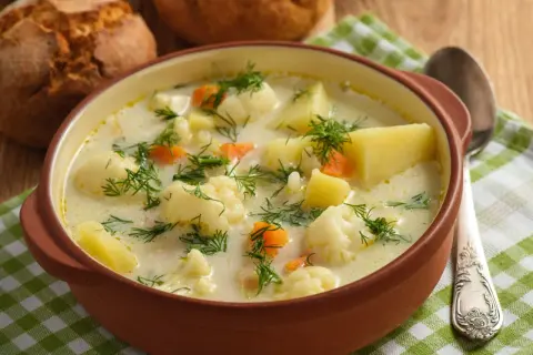 Aksamitna zupa kalafiorowa jak u babci, z ziemniakami i marchewką, posypana koperkiem. Obok bułeczki i łyżka.