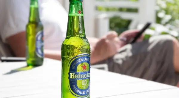 Orzeźwiająca butelka Heineken 0.0 procent, idealna na letni relaks. W tle osoba korzystająca z telefonu.