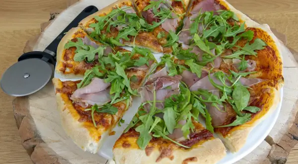 Pyszne pomysły na pizzę: cienkie ciasto, sos pomidorowy, ser, szynka i rukola. Obok leży nóż do pizzy.