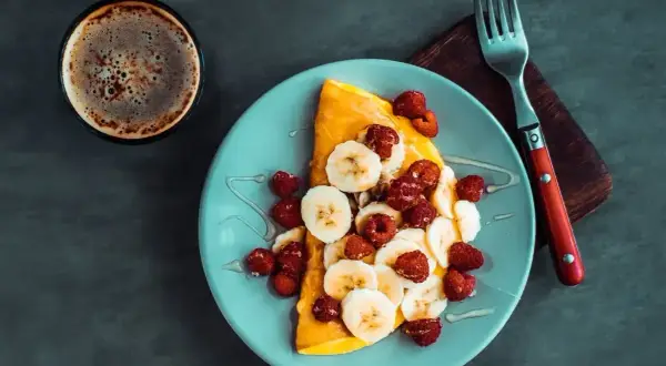 Słodki omlet z bananami i malinami, polany syropem, obok filiżanka kawy. Idealny na słodki początek dnia.