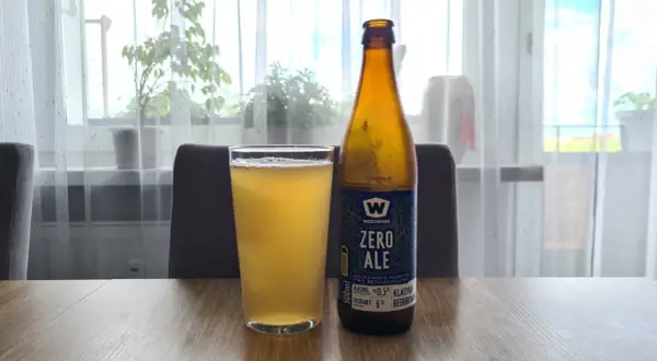 Szklanka i butelka piwa zero na drewnianym stole. Za nimi okno z firanką i roślinami.