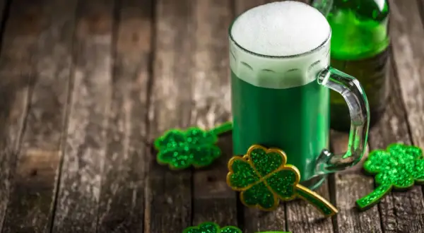 Zielone piwo irlandzkie z pianką, butelka i błyszczące koniczynki na drewnianym stole.