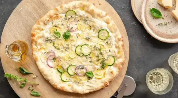 Pyszna pizza bez sosu pomidorowego z cukinią, cebulą i ziołami, podana na drewnianej desce.