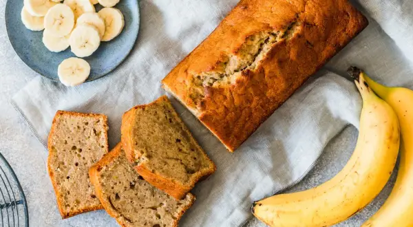 Pyszny chlebek bananowy z dwóch bananów, pokrojony na kromki. Obok leżą świeże banany i talerzyk z plasterkami.