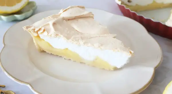 Pyszna tarta cytrynowa z puszystą bezą na wierzchu, podana na białym talerzu. Obok leży połówka cytryny.