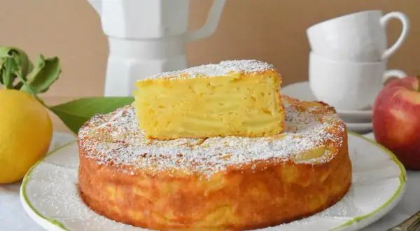 Pyszna torta di mele, posypana cukrem pudrem, z kawałkiem jabłka na wierzchu. Obok cytryna i jabłko.