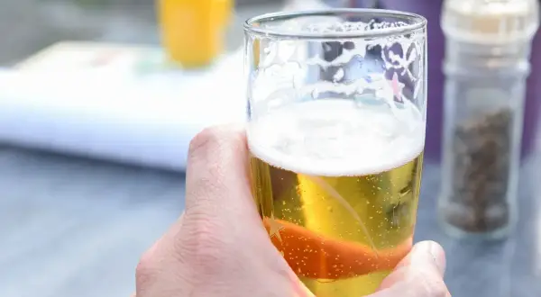 Dłoń trzymająca szklankę orzeźwiającego piwa dietetycznego. W tle widać pomarańczowy napój i młynki do przypraw.