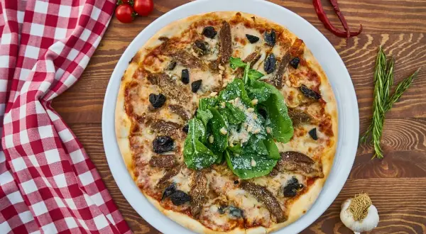 Idealna pizza z piwem na drewnianym stole, ozdobiona rukolą i parmezanem. Obok czosnek, pomidorki i rozmaryn.