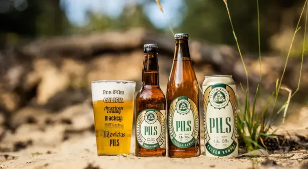 Szklanka, butelka i puszka piwa lager "Pils" od Trzech Kumpli na piasku, wśród traw.