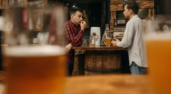 Dwóch mężczyzn rozmawia przy barze, zastanawiając się, ile węglowodanów ma piwo. Na stole stoją kufle i beczka.
