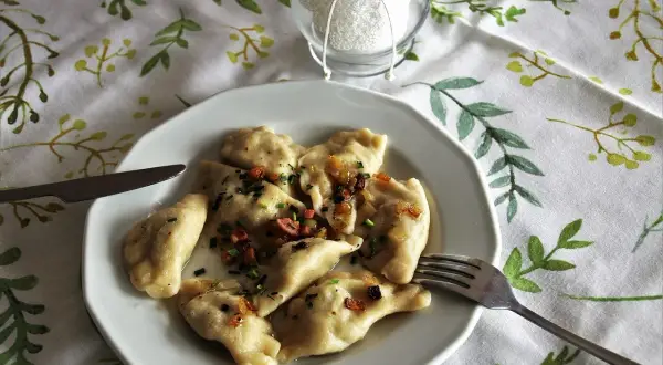 Pyszne pierogi Ania gotuje, polane skwarkami i szczypiorkiem. Na talerzu obok widelca i noża.