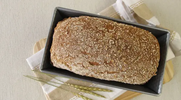Świeżo upieczony chleb orkiszowy w keksówce, posypany ziarnami. Obok leżą kłosy zbóż i lniana ściereczka.