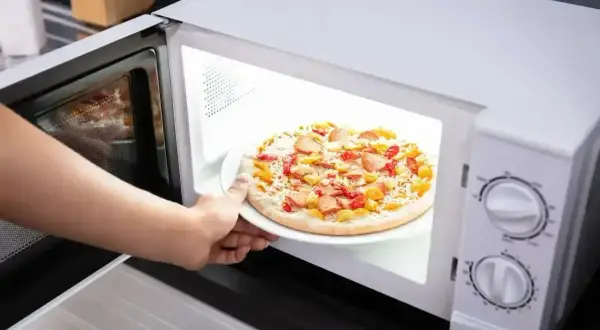 Ręka wkłada do mikrofali talerz z pizzą Giusseppe. Szybkie danie gotowe do podgrzania.