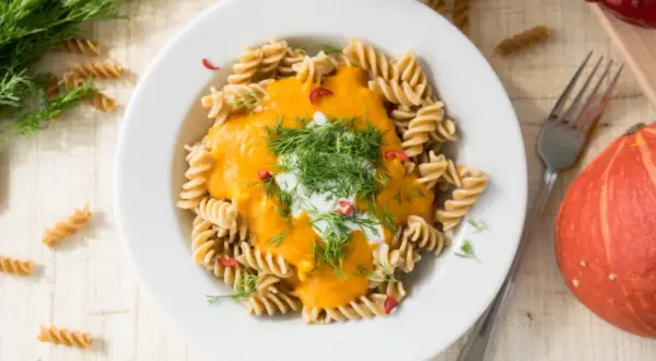 Makaron fusilli z kremowym sosem z **hokkaido dyni**, udekorowany koperkiem i chili. Obok talerza leży cała dynia.
