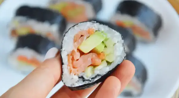 Ręka trzyma kawałek sushi z łososiem i awokado w środku. Na talerzu widać więcej takich roladek.